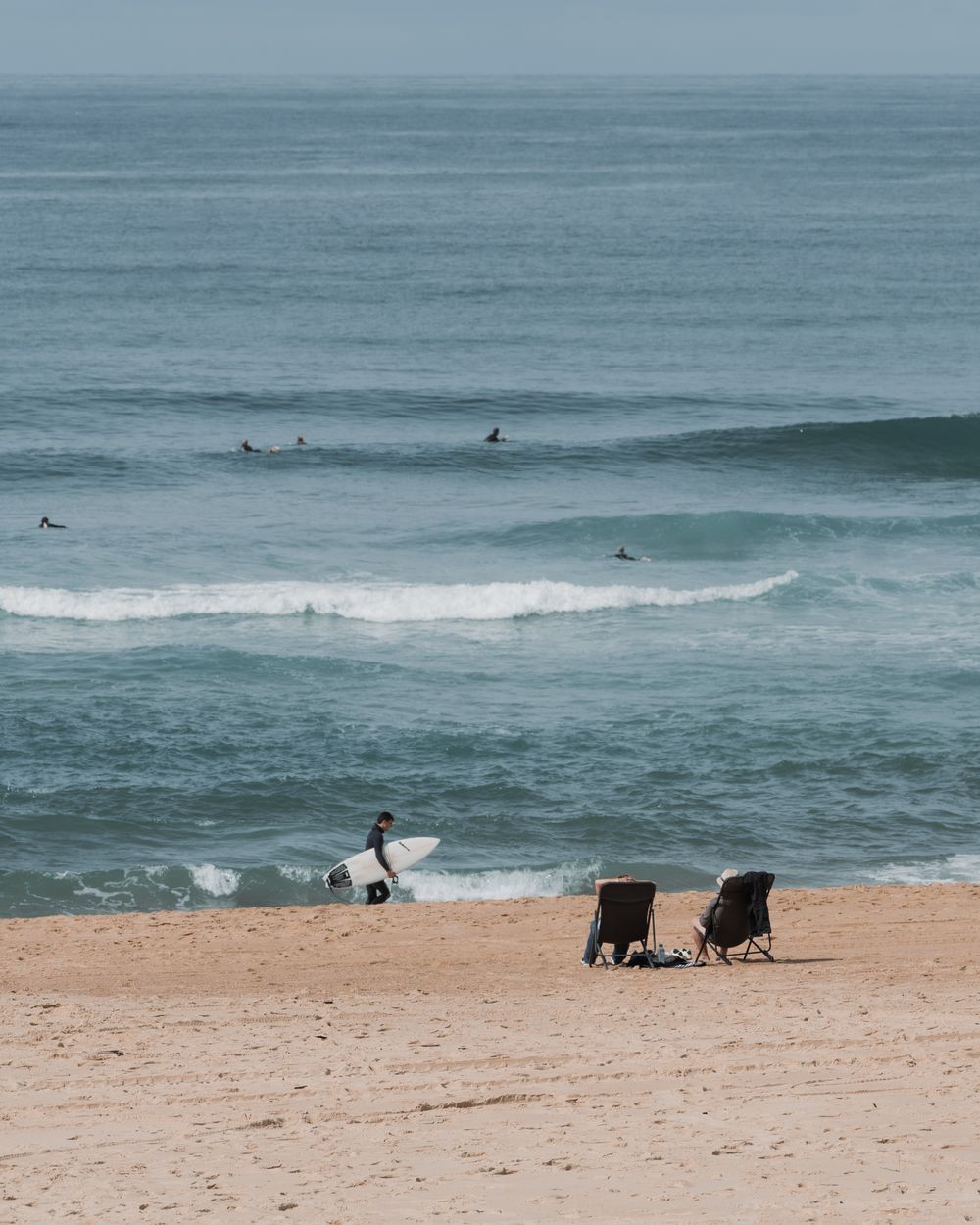 A surfer walking along the sandy beach of Hossegor, France, with waves rolling in