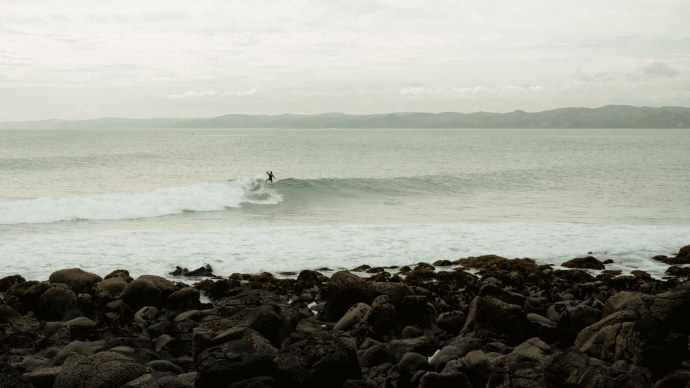 A surfer riding a wave along the rocky, hill-backed coastline of Raglan, New Zealand